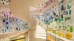 A glowing white spiral staircase inside La Galerie Dior in Paris, encased by glass displays filled with miniature color-sorted dresses, handbags, and shoes. See on the Galerie Dior Tour